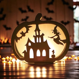 Könnte beinhalten: Eine weiße, hölzerne Halloween-Dekoration in Form eines Kürbisses mit einem ausgeschnittenen Design eines Spukhauses, einer Hexe, die auf einem Besen fliegt, und einem Kessel. Die Dekoration wird von innen beleuchtet und erzeugt ein warmes Leuchten.