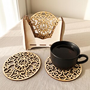 2 wooden coasters are placed on the table. A cup of tea is placed on 1 coaster. There is a coaster holder with coasters inside. Placemats are cut out with floral motifs.