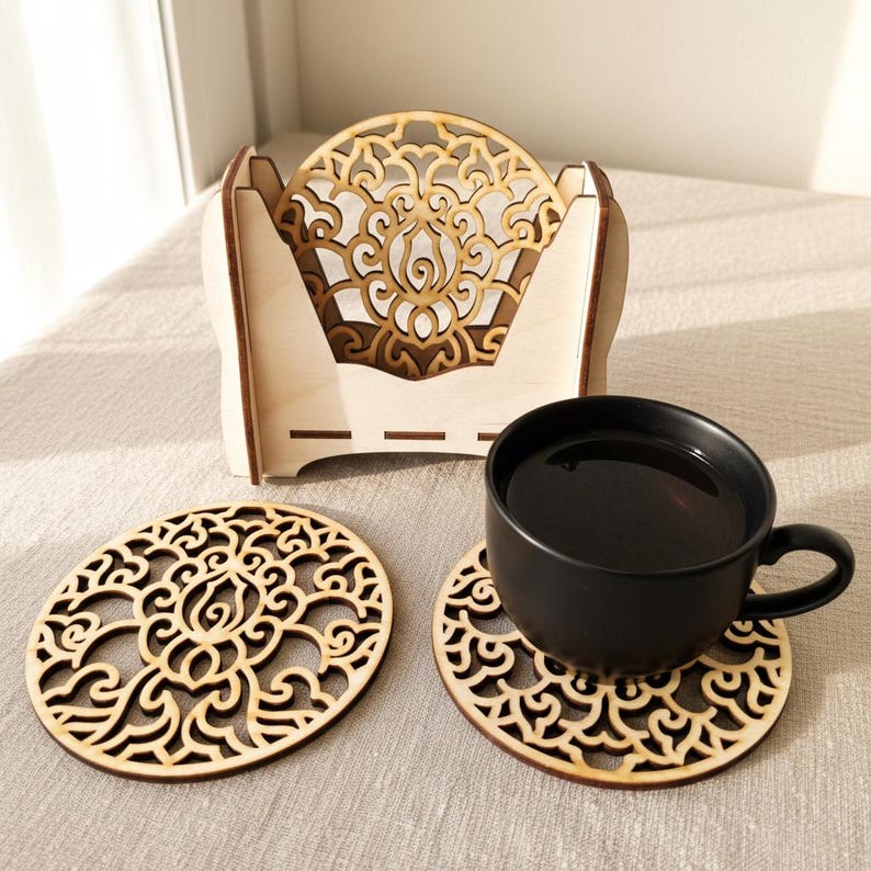 2 wooden coasters are placed on the table. A cup of tea is placed on 1 coaster. There is a coaster holder with coasters inside. Placemats are cut out with floral motifs.