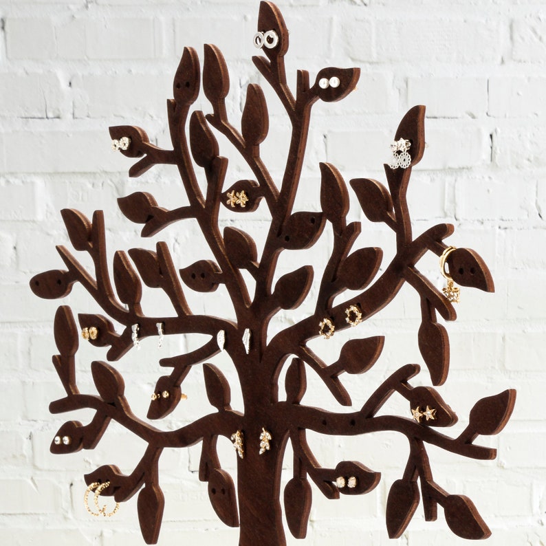 Close-up of the tree. Dark brown laser cut wooden jewelry holder. The tree has holes for earrings. Other jewelry can be hung on the branches. Several pairs of earrings are hung on the tree.