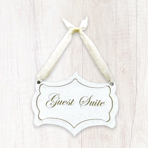May include: White wooden sign with a gold ribbon hanging from it. The sign says "Guest Suite" in gold lettering.