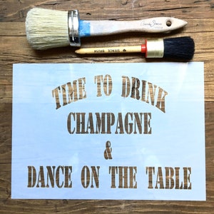 May include: A white stencil with the text "Time to Drink Champagne & Dance on the Table". The stencil is on a wooden surface with two paint brushes.