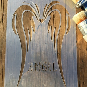 May include: A clear plastic stencil featuring angel wings and the word "ANGEL" in a decorative font. The stencil is placed on a wooden surface, with paint brushes visible in the background. The design includes a heart shape.
