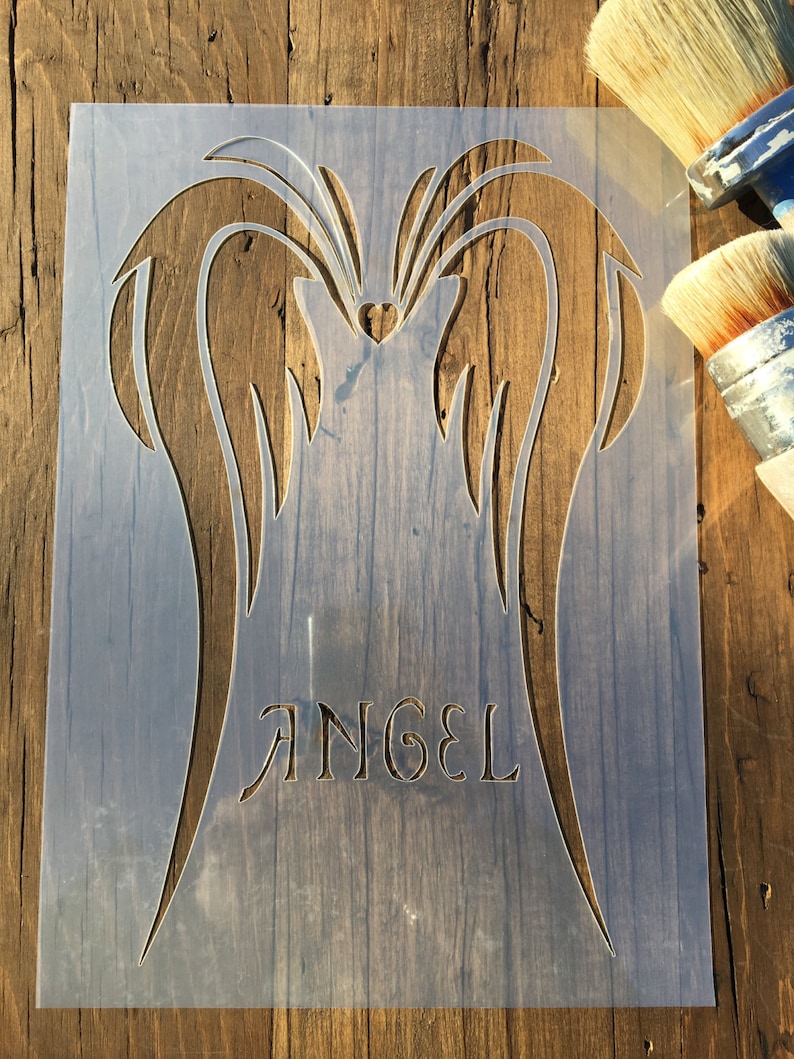 May include: A clear plastic stencil featuring angel wings and the word "ANGEL" in a decorative font. The stencil is placed on a wooden surface, with paint brushes visible in the background. The design includes a heart shape.