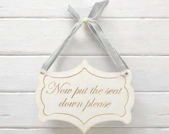 May include: White wooden sign with a decorative border and a gold inscription that reads "Now put the seat down please". The sign is hanging from a white wall with a gray ribbon.