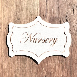 Puede incluir: Un letrero de madera blanco con un borde decorativo y la palabra "Nursery" escrita en cursiva.