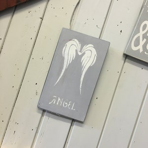 May include: A gray wooden sign with white angel wings and the word "ANGEL" stenciled on it. The sign is rectangular and has a distressed look. The background is a white wooden wall.