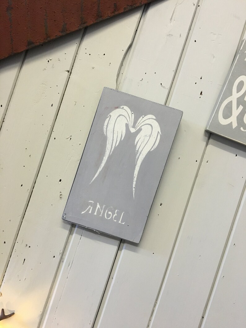 May include: A gray wooden sign with white angel wings and the word "ANGEL" stenciled on it. The sign is rectangular and has a distressed look. The background is a white wooden wall.
