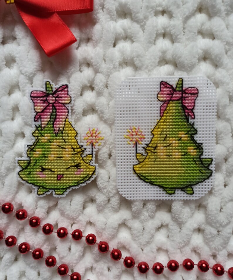 May include: Two green Christmas tree ornaments with pink bows and yellow lights. Each tree has a pink bow and a sparkler. The ornaments are on a white fluffy background.