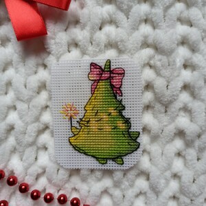 May include: A green Christmas tree with a pink bow and a sparkler, cross-stitched on white Aida cloth.