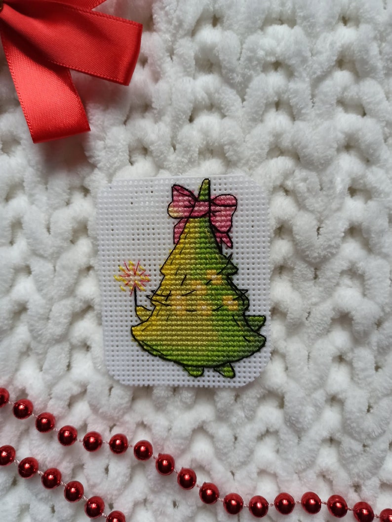 May include: A green Christmas tree with a pink bow and a sparkler, cross-stitched on white Aida cloth.