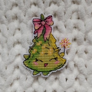 May include: A green and yellow Christmas tree with a pink bow and a sparkler. The tree has a smiling face and is embroidered.