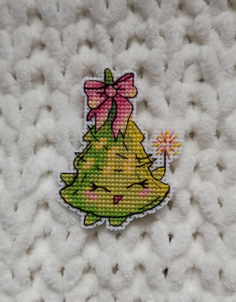 May include: A green and yellow Christmas tree with a pink bow and a sparkler. The tree has a smiling face and is embroidered.