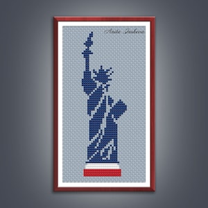 Peut inclure: Une broderie au point de croix encadrée de la Statue de la Liberté en bleu marine sur un fond gris clair. La base présente un motif rouge, blanc et bleu. Le nom de l'artiste, "Aaila Iasheva", est en haut à droite.