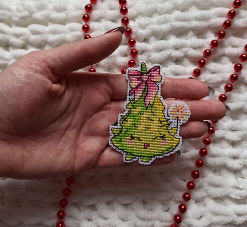 May include: A hand holding a green and yellow cross-stitch Christmas tree ornament with a pink bow and a yellow star on top.