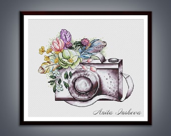 May include: A vintage camera with a floral design, featuring pink, yellow, and green flowers. The camera is surrounded by a bouquet of flowers and leaves. The text "Anita Insheva" is written in cursive at the bottom of the image.