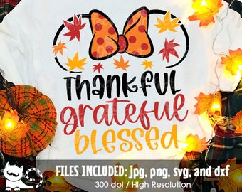 May include: A white t-shirt featuring a graphic of a polka-dot orange bow with mouse ears, surrounded by red and yellow autumn leaves. The text reads Thankful in black, grateful in red, and blessed in yellow, with file format details below.