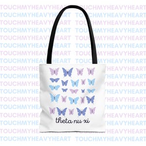 Op de afbeelding: Witte tote bag met zwarte handvatten met een patroon van blauwe en paarse vlinders. De tekst "theta nu xi" is in zwart onderaan de tas gedrukt.