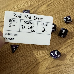 May include: A white film slate with the text "Roll the Dice" and other handwritten details, surrounded by several purple and black dice. The dice have white numbers. The slate reads "Roll 1, Scene Dice, Take 2."