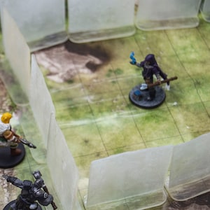 May include: A tabletop game scene featuring four miniature figures, including a warrior with a sword, a wizard with a staff, and a warrior with a shield. The figures are placed on a green and brown gridded game board with clear plastic walls surrounding them.