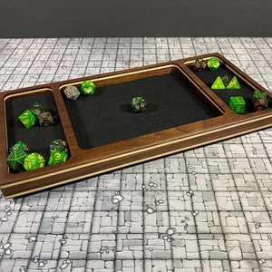 May include: A wooden dice tray with a black felt lining and multiple compartments for dice. The tray has a light wood border and a dark wood centre. There are several green and gold dice in the tray.