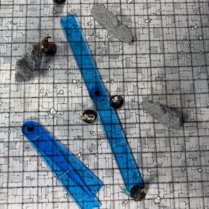 May include: A tabletop role-playing game setup with a gray stone-like surface. Two blue plastic templates are positioned diagonally. Miniature figures and terrain pieces are scattered across the surface, creating a scene for a fantasy adventure.