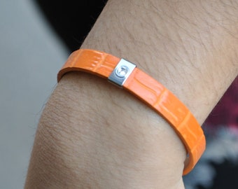 May include: A bright orange bracelet with a textured, embossed surface resembling reptile skin. The band features a central rectangular silver-toned metal accent piece that includes a small, circular embedded clear stone or crystal detail.