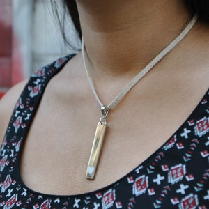 May include: A silver-toned vertical bar pendant necklace featuring a small cutout heart shape at the bottom. The pendant hangs from a light beige, textured cord. The necklace is worn against a black sleeveless top with a geometric red and white pattern.