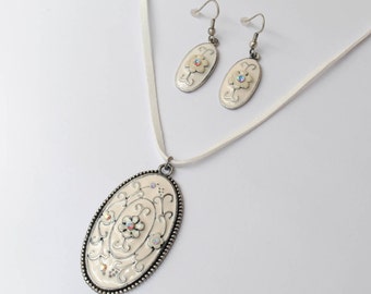 May include: A cream-colored jewelry set featuring an oval pendant necklace and matching earrings. The pendant and earrings have a floral design with iridescent accents and a silver-tone border. The necklace has a white cord.