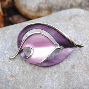 Könnte beinhalten: Eine silberfarbene Brosche mit einem Design aus lila und rosa Emaille. Die Brosche ist blattförmig mit einem geschwungenen, abstrakten Design.