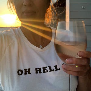 Könnte beinhalten: Weißes T-Shirt mit dem Text "OH HELL" in Schwarz, kombiniert mit einem Weinglas. Der Wein hat eine hellrosa Farbe. Das Bild wurde bei Sonnenuntergang aufgenommen, wobei die Sonnenstrahlen durchscheinen.