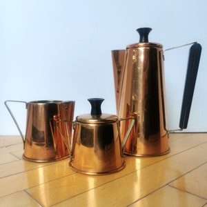 May include: A copper coffee pot, creamer and sugar bowl set. The coffee pot has a black handle and a black knob on the lid. The creamer and sugar bowl have black knobs on the lids.