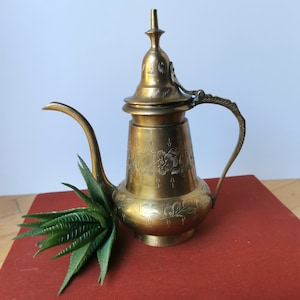 May include: A gold-colored brass teapot with an ornate design and a lid. The teapot is sitting on a red book.