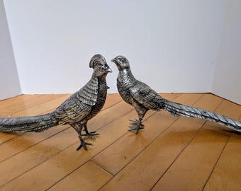 May include: Two silver-colored pheasant figurines. The birds have detailed feather textures and long, flowing tails. One pheasant has a distinctive crest. They are standing on a wooden surface, creating a decorative display.