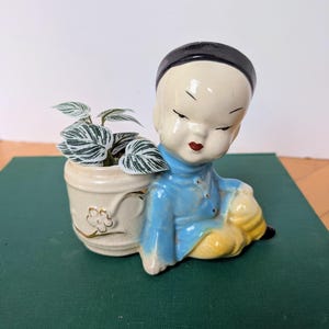 May include: A vintage ceramic planter in the shape of a seated child. The figure wears a blue jacket, yellow pants, and a black cap. The planter is a cream color with a floral design and holds a small plant with green and white striped leaves.