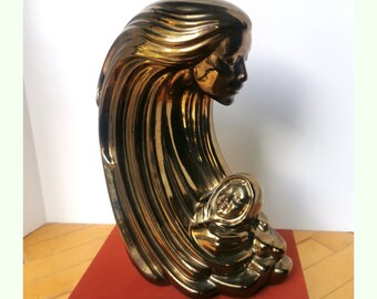 May include: A gold-toned ceramic sculpture of a woman with flowing hair cradling a child. The sculpture is abstract and stylized, with the woman's hair forming a wave-like shape around the child.