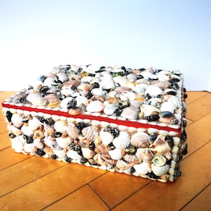 May include: A square box covered in a mosaic of small seashells. The box has a red trim around the top edge.