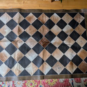May include: A black and brown checkered rug with a diamond pattern. The rug is made of leather and has a fringed border.