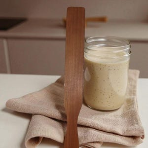 May include: A wooden spatula stands beside a glass jar containing a creamy, speckled mixture. The jar sits on a folded, light brown cloth. The spatula and jar are on a white surface, with a blurred kitchen background.