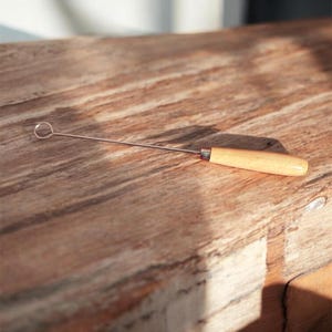 Pode incluir: Uma ferramenta de aço inoxidável com um pequeno laço em uma extremidade e uma alça de madeira clara. A ferramenta está sobre uma superfície de madeira com grão natural.