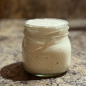 May include: A glass jar filled with a light brown, bubbly sourdough starter.