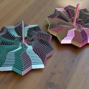 Puede incluir: Dos esculturas geométricas abstractas en tonos verdes, morados y rosas sobre una superficie de madera. Cada escultura tiene una varilla central y un diseño en capas en forma de estrella. Las esculturas son objetos decorativos.