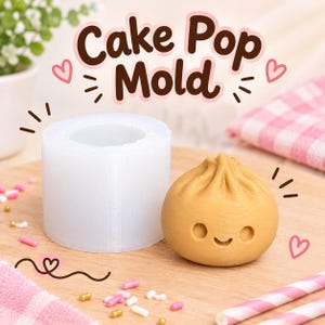 Puede incluir: Un molde de silicona cilíndrico y translúcido para cake pops junto a un cake pop de color canela, redondo y con forma de dumpling que tiene una cara sonriente. El texto "Cake Pop Mold" aparece en marrón arriba, con corazones y rayas rosas.