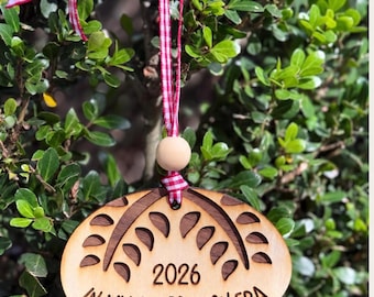 May include: A light-colored wooden oval ornament featuring a laser-engraved design of wheat stalks, the year 2026, and the text IN MY SOURDOUGH ERA. It hangs from a red and white checkered ribbon with a small, round, natural wooden bead.