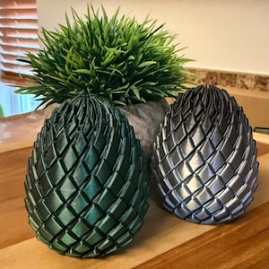 Puede incluir: Dos esculturas decorativas en forma de huevo con una textura tejida. Una es verde metálico, la otra plateada metálica. Se encuentran sobre una superficie de madera, con una pequeña planta verde en una maceta de piedra detrás de ellas. Los huevos son probablemente objetos decorativos.