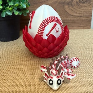 Puede incluir: Un huevo decorativo con temática de béisbol con costuras rojas y detalles de bate de béisbol, encajado en un soporte rojo en forma de pétalo. También hay una pequeña figura de dragón de béisbol blanco y rojo.