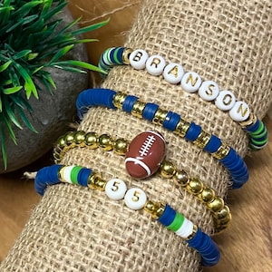 May include: A set of four bracelets featuring blue and green beads, gold beads, a white football charm, and white letter beads spelling "BRANSON" and "55".
