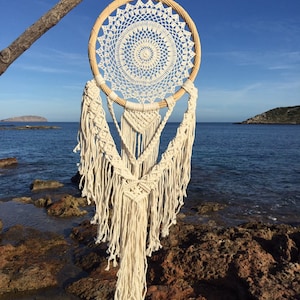 Op de afbeelding: Een witte gehaakte droomvanger met een houten ring en witte franjes die hangt aan een tak tegen een blauwe lucht en oceaan achtergrond.