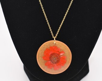 May include: A gold-colored chain necklace with a circular pendant. The pendant features a bright red flower encased in a clear resin. The flower has a dark center and is set against a light brown background.