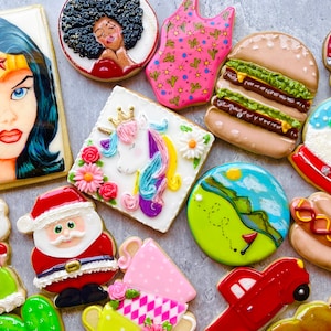 May include: A collection of decorated sugar cookies featuring various themes, including a superhero, a unicorn, a snowman, a hamburger, a hot dog, a camper, a golf course, a Christmas tree, a Santa Claus, and a cactus.